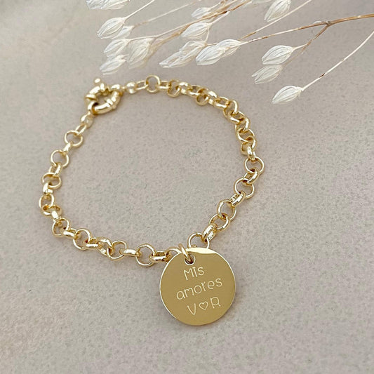 Pulsera formada por una cadena de color dorado con un dije de color dorado que tiene una frase grabada en su superficie. 