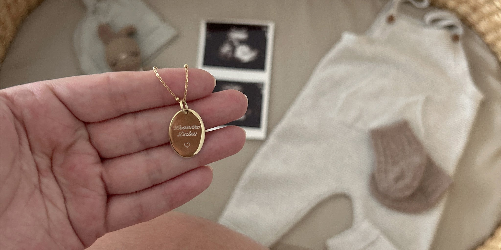 Una mano sujeta un collar dorado con un dije dorado que tiene un nombre y un corazón grabados en su superficie. En el fondo se ve una cuna y sobre esta hay unas medias de bebe, un overol de bebe y una ecografía.