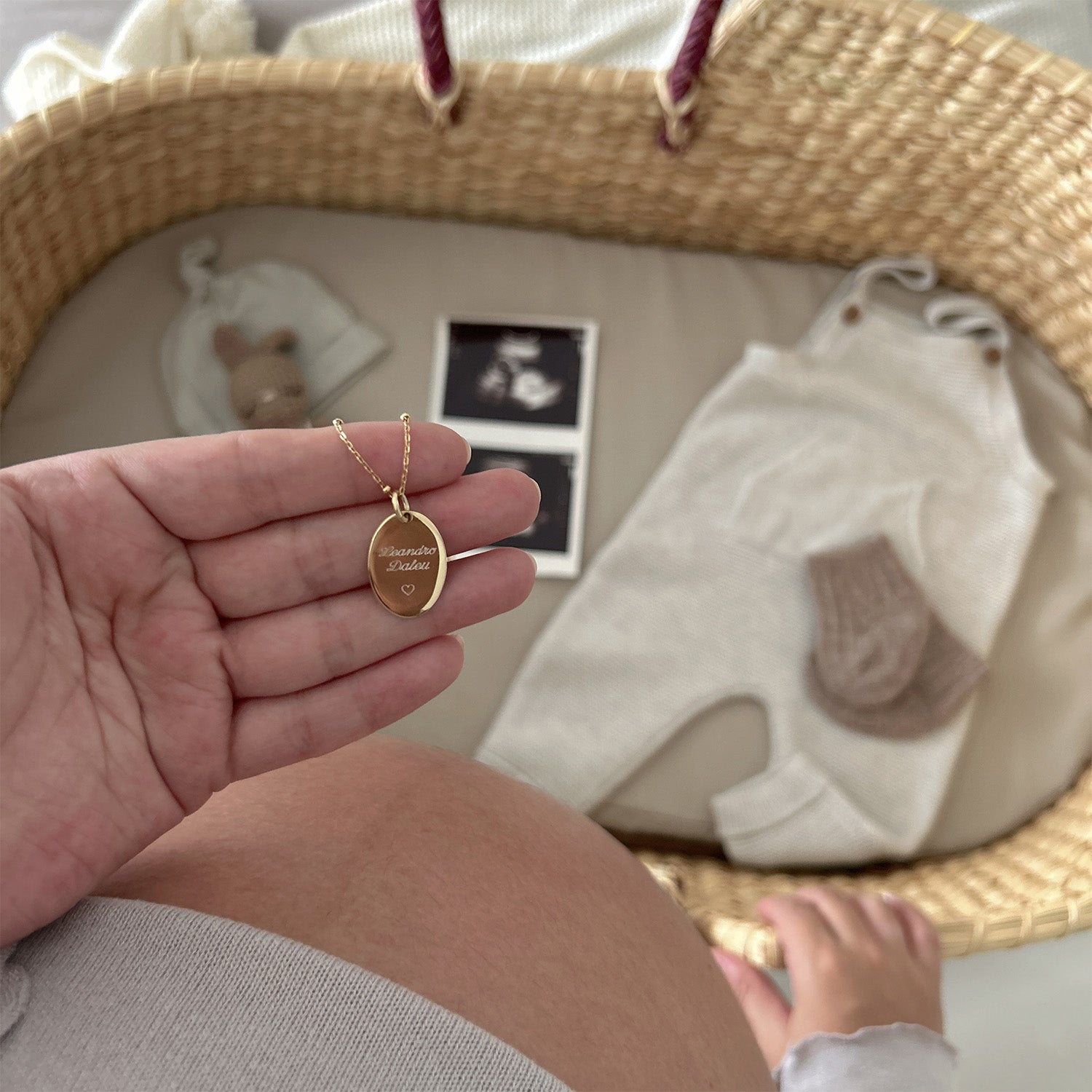 Una mano sujeta un collar dorado con un dije dorado que tiene un nombre y un corazón grabados en su superficie. En el fondo se ve una cuna y sobre esta hay unas medias de bebe, un overol de bebe y una ecografía.